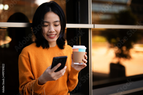 Pretty chinese girl using mobile phone and holding coffee to go. Beautiful woman enjog in coffee while walking on the city street.