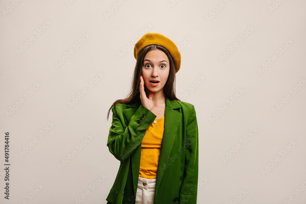 Young fair-skinned woman brunette with open mouth looking at camera in amazement on white background. Portrait of lady in beret, blouse, jacket and pants in studio. Emotional reaction concept Young fair-skinned woman brunette with open mouth looking at camera in amazement on white background. Portrait of lady in beret, blouse, jacket and pants in studio. Emotional reaction concept