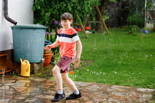 School kid boy running through heavy summer rain in garden. Happy smiling wet child having fun with splashing and jumpin in puddles. Activity for children on rainy weather day. photo