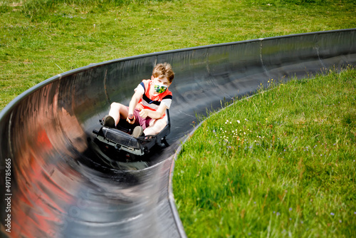 Young school kid boy having fun riding summer toboggan run sled down a hill in Hoherodskopf, Germany. Active child with medical mask making funny activity otudoors. Family leisure with kids. photo