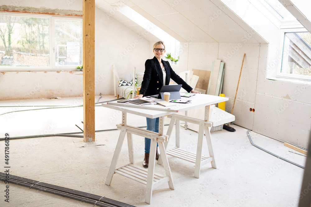 pretty young architect working on construction site in a loft and standing by a high table wearing glasses and a black jacket