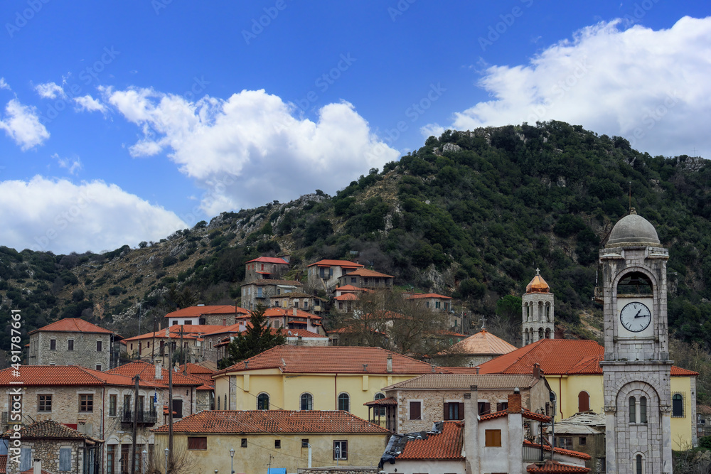 Greek village with traditional low-rise houses against blue sky with clouds. Day view of historic settlement with red roof tiles residences and church bell tower in Dimitsana, Arcadia Peloponnese.