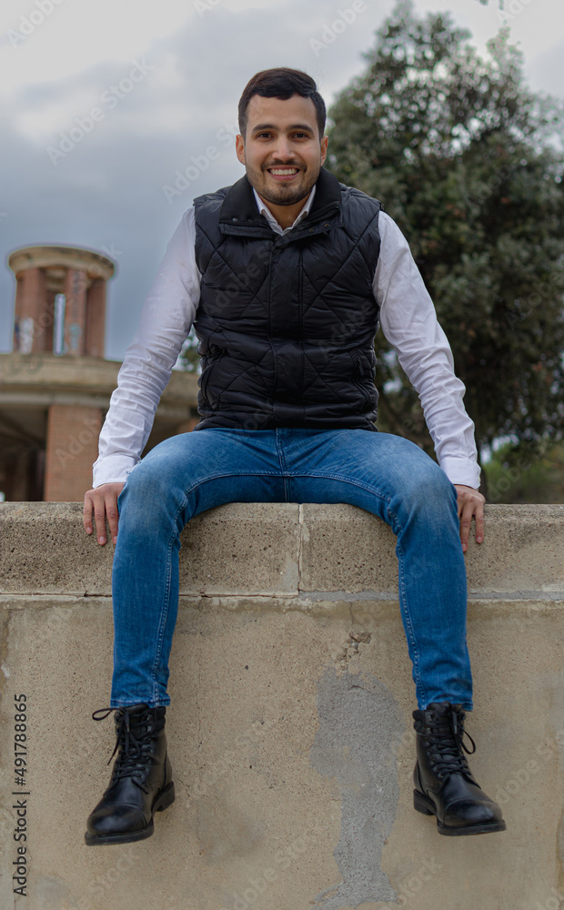 Full length image of a handsome Latino man sitting on the top of a wall on a cloudy day with an unfocused background composed of a tree and a brick tower behind him. Full length image of a handsome Latino man sitting on the top of a wall on a cloudy day with an unfocused background composed of a tree and a brick tower behind him.