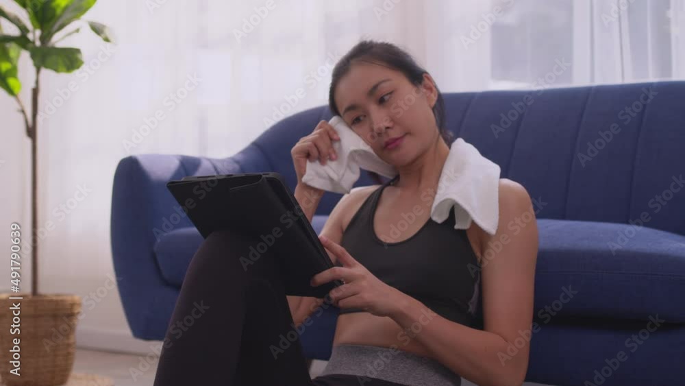 Asian woman in sportswear wiping forehead with towel and using digital tablet while resting after workout in home. Asian woman in sportswear wiping forehead with towel and using digital tablet while resting after workout in home.