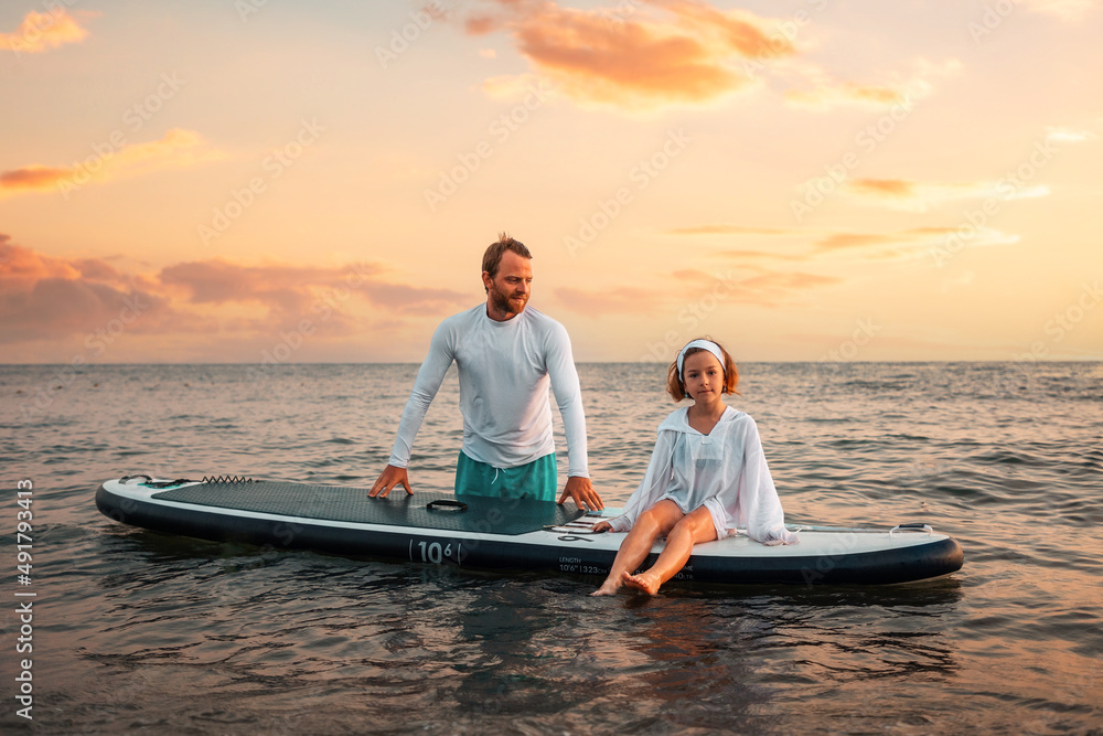 Father and daughter surfing on a sup board. Sea and sunset sky at the background. Summer vacations. Copy space