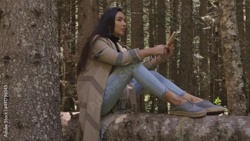 pretty woman using phone in forest nature. Beautiful woman relax in forest and browsing smartphone outdoors. Pensive woman sitting on wooden log. Summer forest background. High quality 4k footage