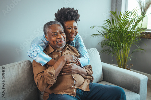 Loving senior man embracing his daughter while sitting on sofa smiling. An attractive young woman embracing her grandfather from back. 