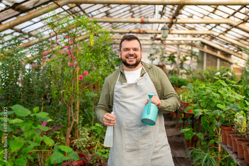 Obraz premium Portrait of a young smiley man in a greenhouse. He looks into the camera, smiles and holds a watering can in his hands.