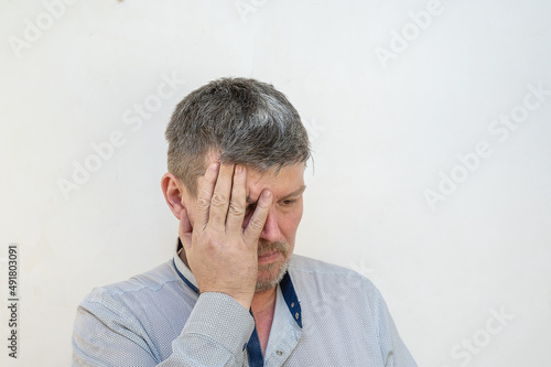 A mature man covers his face with his hand against the light wall. A male with a short haircut and graying hair The emotion of grief, of loss