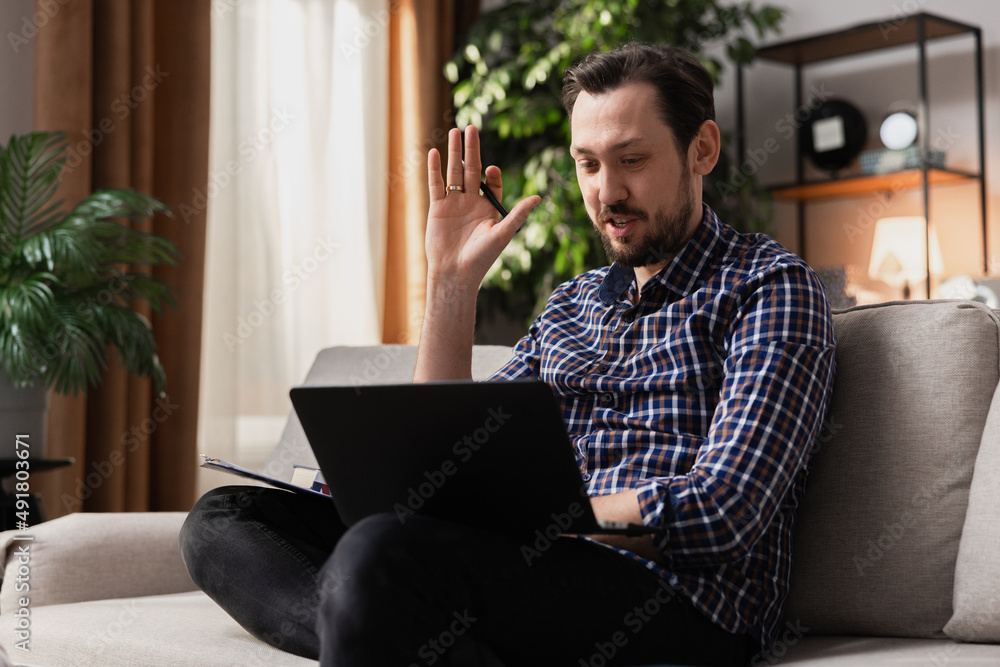 A final year student is chatting with friends on a laptop. A bearded middle-aged man sits on the living room couch and waves his hand in greeting during a video chat. A final year student is chatting with friends on a laptop. A bearded middle-aged man sits on the living room couch and waves his hand in greeting during a video chat.