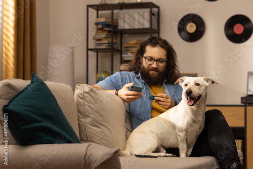 A man sits on a couch in his living room and pays with a credit card at an online store. The boy enters the credit card number on a smartphone accompanied by his dog.