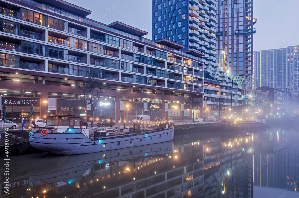 Fototapeta premium Rotterdam, The Netherlands, January 12, 2022: modern residential buildings, partly under construction, and historic vessels reflect in the still water of Wijnhaven harbour on a misty morning