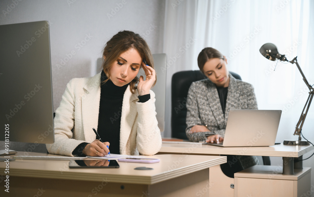 Two young adult women work in an office or coworking space on computers. Two casual businesswomen at work in a modern office space Two young adult women work in an office or coworking space on computers. Two casual businesswomen at work in a modern office space