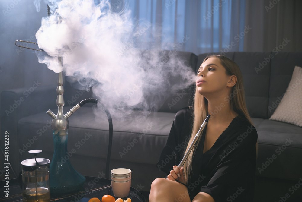 An attractive young adult woman smokes a hookah at home in anticipation of a party with her friends