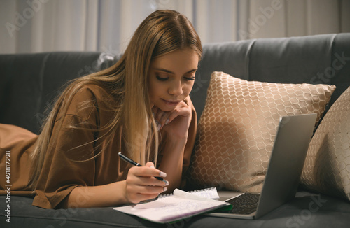 A teenager girl is engaged in homeschooling using a laptop  making notes in a notebook. Study on the couch  distance learning and work