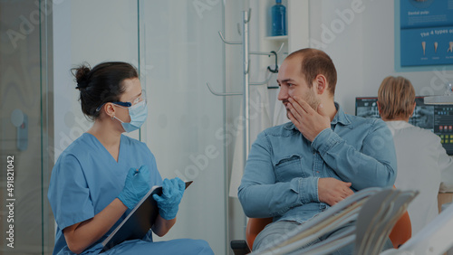 Patient explaining toothache to dentistry nurse at stomatology visit. Orthodontic assistant taking notes to prepare for oral care procedure to cure caries pain with dental tools in cabinet.
