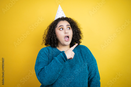Young beautiful woman wearing a birthday hat over isolated yellow background amazed and pointing with hand and finger to the side © Irene