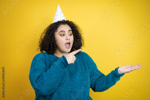 Young beautiful woman wearing a birthday hat over isolated yellow background surprised, showing and pointing something that is on her hand © Irene