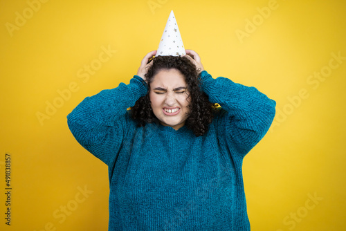 Young beautiful woman wearing a birthday hat over isolated yellow background suffering from headache desperate and stressed because pain and migraine with her hands on head © Irene