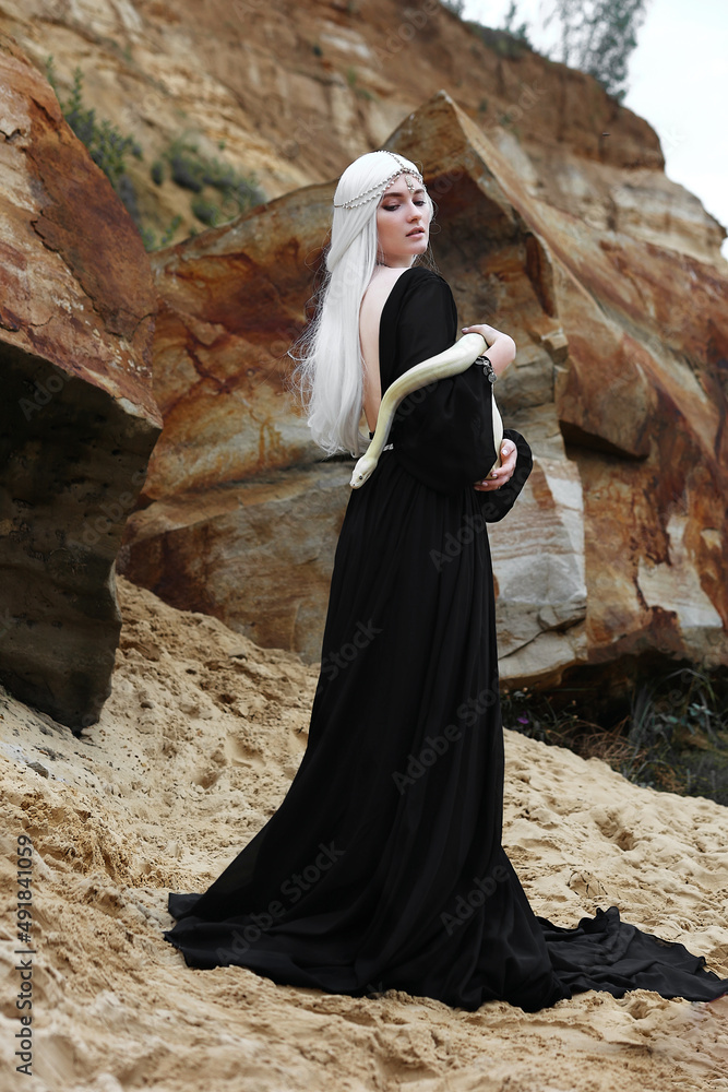 Beautiful white haired girl in long black dress with white snake among rocks and sand
