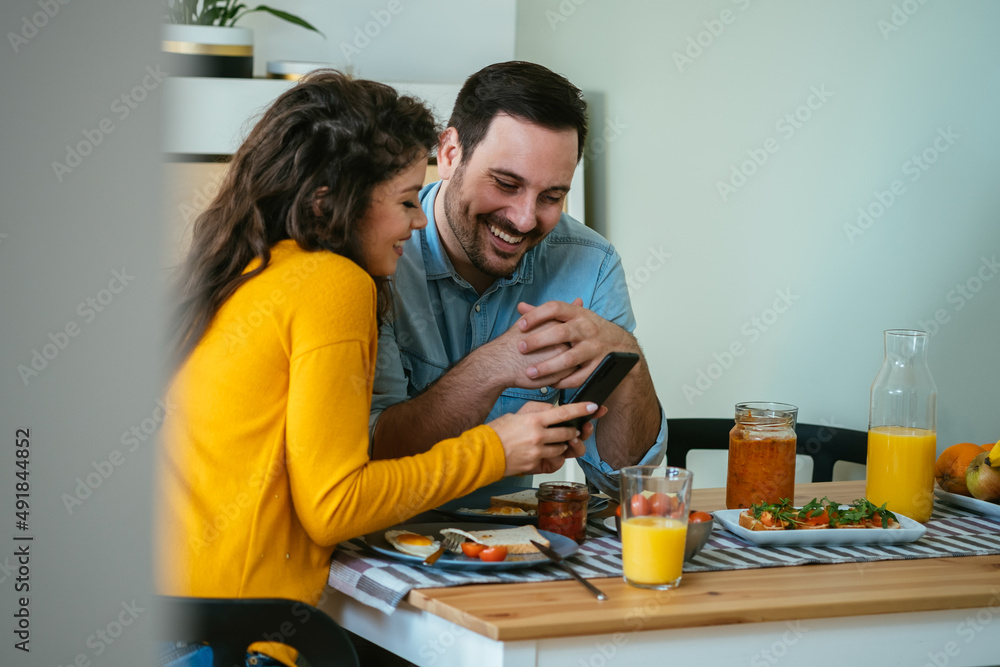 Happy Couple Using Mobile Phone Together While Eating Breakfast at Home Happy Couple Using Mobile Phone Together While Eating Breakfast at Home