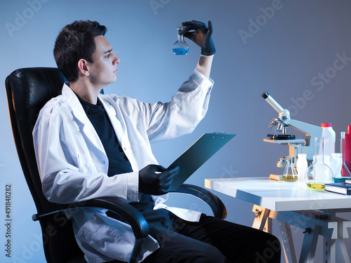 chemist is sitting at table. Man chemist is holding test tube. Laboratory technician with clipboard in his hands. Table with microscope and test tubes in front of scientist. Man chemist career