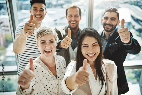 Their success rate is very high. Portrait of a group of cheerful businesspeople standing together while showing thumbs up to the camera inside of the office.