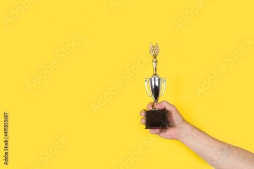 Photography Mano de hombre sosteniendo y levantado un trofeo sobre un fondo amarillo liso y aislado