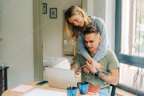 Young attractive blonde hugging her boyfriend while he is working on a laptop.