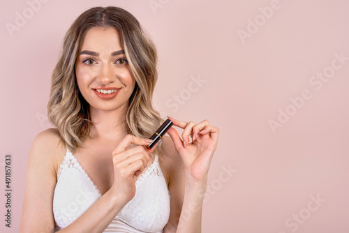 Beutiful blonde girl with natural makeup on light pink background, holding a lipstick , copy space
