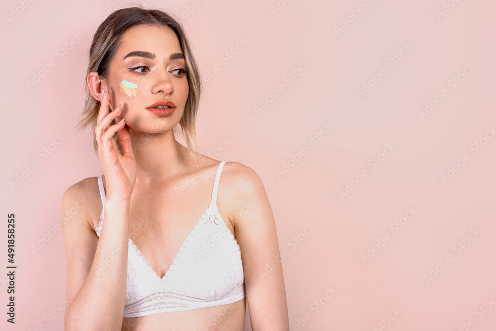 Beautiful blonde girl with natural makeup on light pink background, blending foundation on her face, copy space Beautiful blonde girl with natural makeup on light pink background, blending foundation on her face, copy space