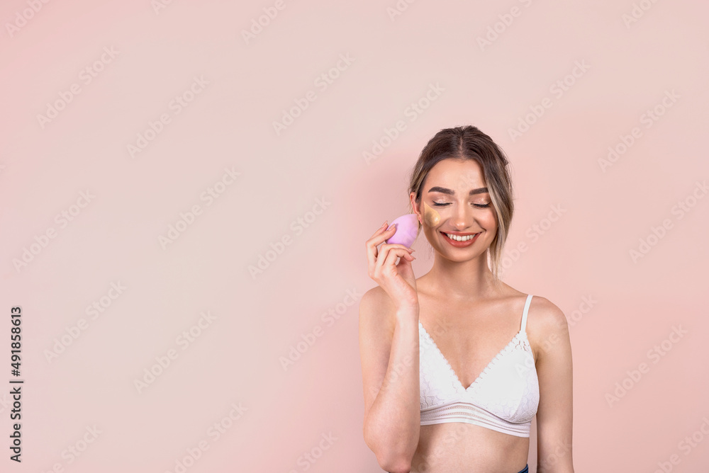 Fototapeta premium Beautiful blonde girl with natural makeup on light pink background, applying foundation on her face with a beauty blender, copy space
