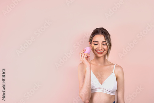 Beautiful blonde girl with natural makeup on light pink background, applying foundation on her face with a beauty blender, copy space © Vita