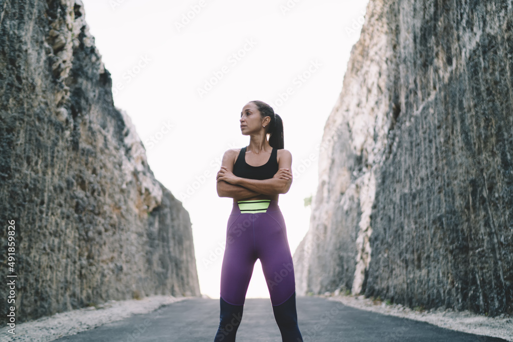 Attractive female runner with crossed hands thinking about sportive endurance and motivation for keeping healthy lifestyle standing at road between rocks, muscular fit girl 20s in tracksuit