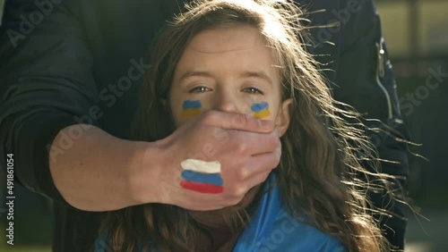 Man's hand with the image of the Russian flag roughly closes the mouth of a little girl with the Ukrainian flag. Children's tears of war. Freedom for Ukraine. Russian aggression.