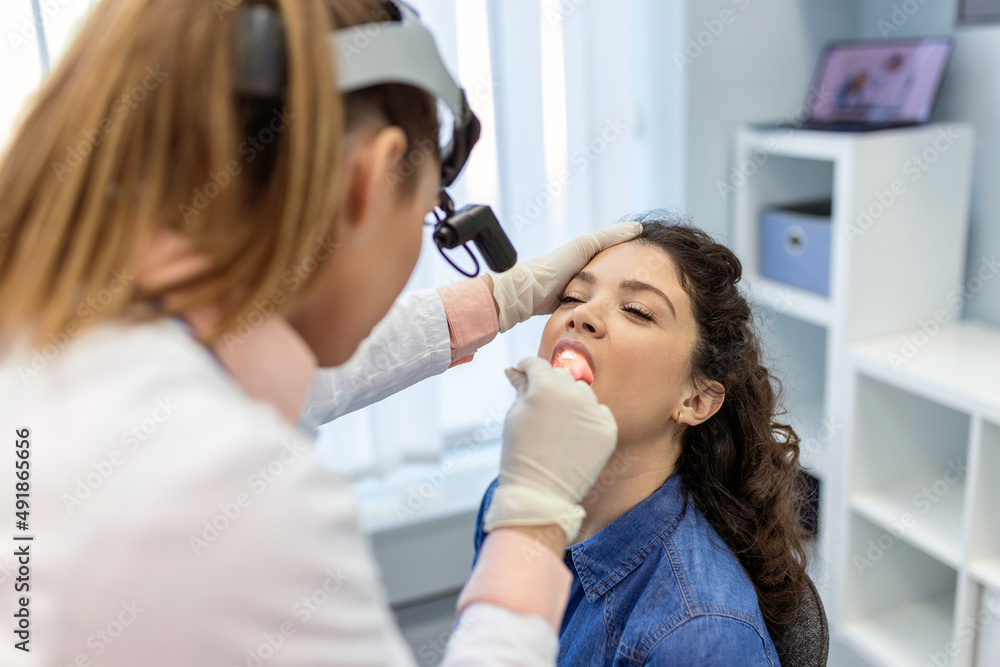 Female patient opening her mouth for the doctor to look in her throat. Female doctor examining sore throat of patient in clinic. Otolaryngologist examines sore throat of patient. Female patient opening her mouth for the doctor to look in her throat. Female doctor examining sore throat of patient in clinic. Otolaryngologist examines sore throat of patient.