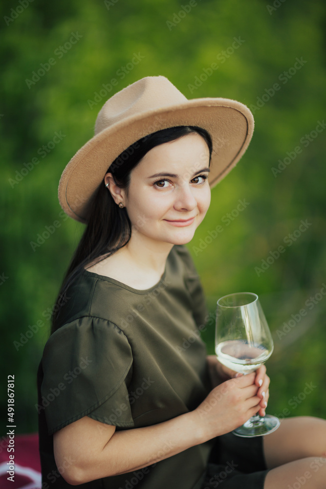 Beautiful happy young woman in green dress and beige hat with glass of wine at nature.