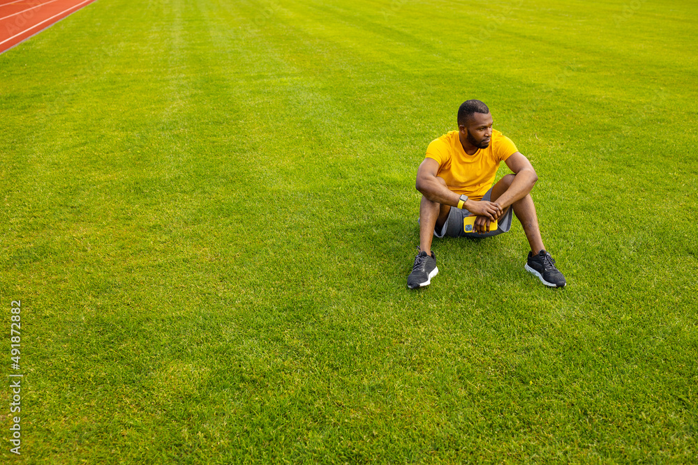 Concentrated athletic man in stylish sportswear sitting at the stadium stretching legs. Sportive african american male working out, doing warm up before training outdoors concept Concentrated athletic man in stylish sportswear sitting at the stadium stretching legs. Sportive african american male working out, doing warm up before training outdoors concept