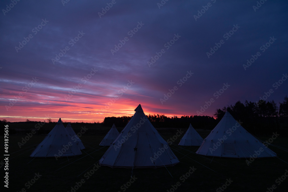 Several white pointed teepees, plunged in darkness, just before sunrise, sleep in a meadow in the English countryside