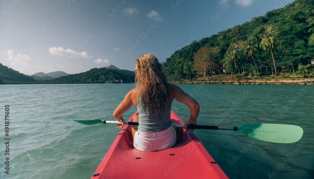 Long haired blonde woman with sunglasses rows bright pink canoe along sea bay water to beach with growing palms. Traveling to tropical countries. Sports girl is sailing on kayak in ocean.