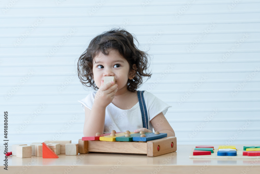 Little cute mixed race kid girl, Caucasian Asian, playing toys build a wooden block, xylophone and colorful puzzle piece on table. White background. Copy space Little cute mixed race kid girl, Caucasian Asian, playing toys build a wooden block, xylophone and colorful puzzle piece on table. White background. Copy space