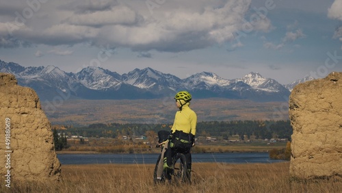 The woman travel on mixed terrain cycle touring with bikepacking. The traveler journey with bicycle bags. Sport bikepacking, bike, sportswear in green black colors. Mountain snow capped, stone arch.