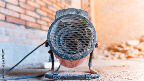 An industrial electric heat gun for space heating stands on the floor against the background of a brick wall.Construction electric heater with a ceramic heating element photo