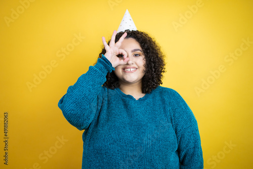 Young beautiful woman wearing a birthday hat over isolated yellow background doing ok gesture shocked with smiling face, eye looking through fingers © Irene