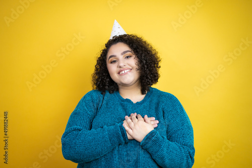 Young beautiful woman wearing a birthday hat over isolated yellow background smiling with her hands on her chest and grateful gesture on her face. © Irene