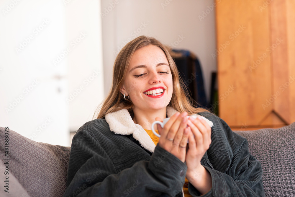Junge Frau mit langen blonden Haaren und Jacke hält eine Tasse auf dem Sofa
Junge Frau mit langen blonden Haaren und Jacke hält eine Tasse auf dem Sofa