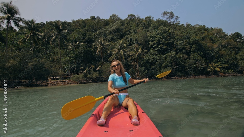 Attractive sportive blonde woman with sunglasses rows pink plastic canoe along sea water against green hills and blue sky. Traveling to tropical countries. Sports girl is sailing on kayak in ocean.