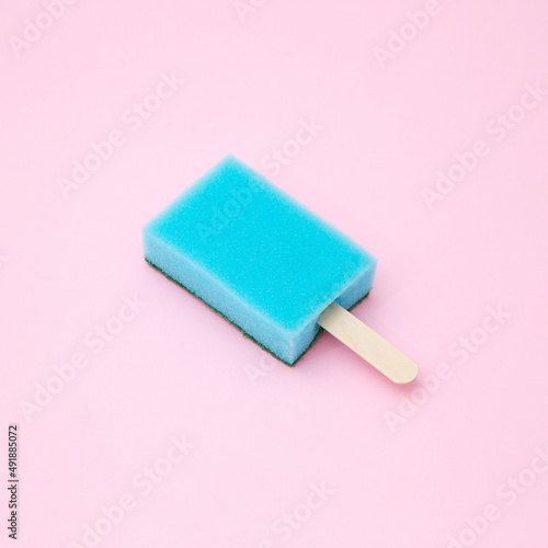 Kitchen sponge on a wooden popsicle stick isolated on a pastel blue background. Creative summer concept. Minimal hygiene or cleaning work idea.