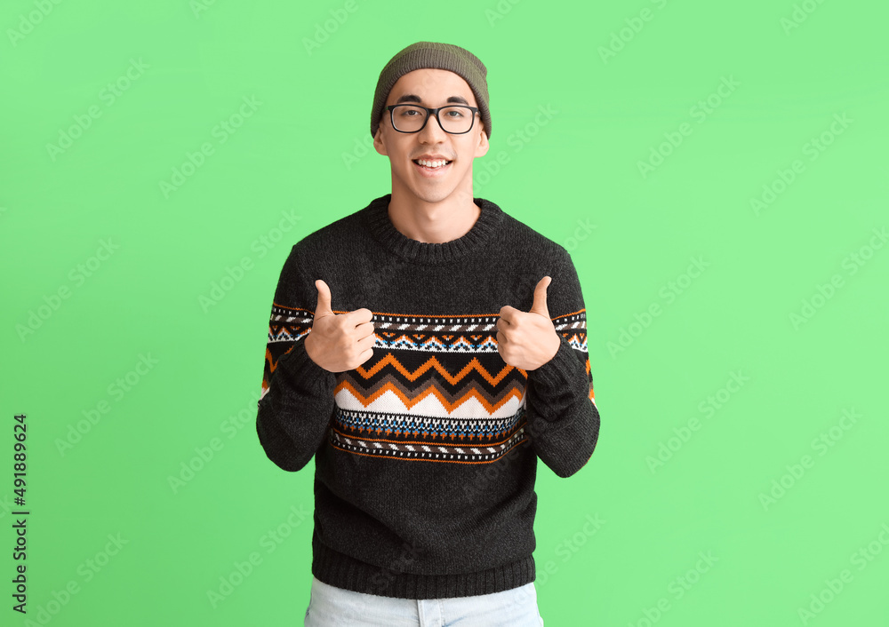 Handsome young Asian man in knitted sweater showing thumb-up on green background Handsome young Asian man in knitted sweater showing thumb-up on green background