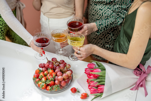 Women clinking glasses with alcohol drinks  celebrating women s day indoors. Close-up on glasses with colorful drinks  bouquet of flowers and fruits on table